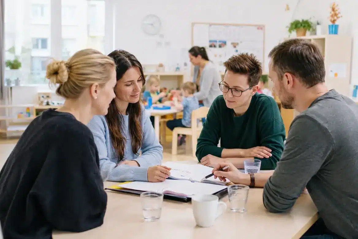 Vier Fachkräfte sitzen an einem Tisch mit Unterlagen und besprechen sich, im Hintergrund spielen Kinder in einer Kita.
